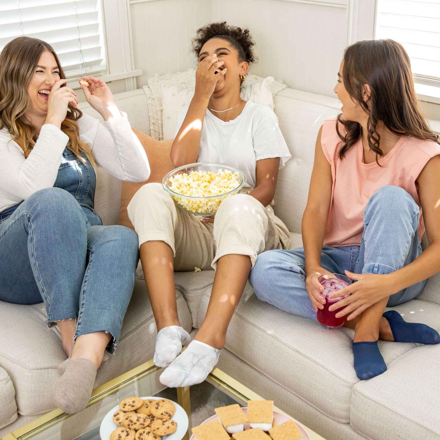 3 mujeres están sentadas en el sofá riendo y comiendo palomitas de maíz mientras usan calcetines