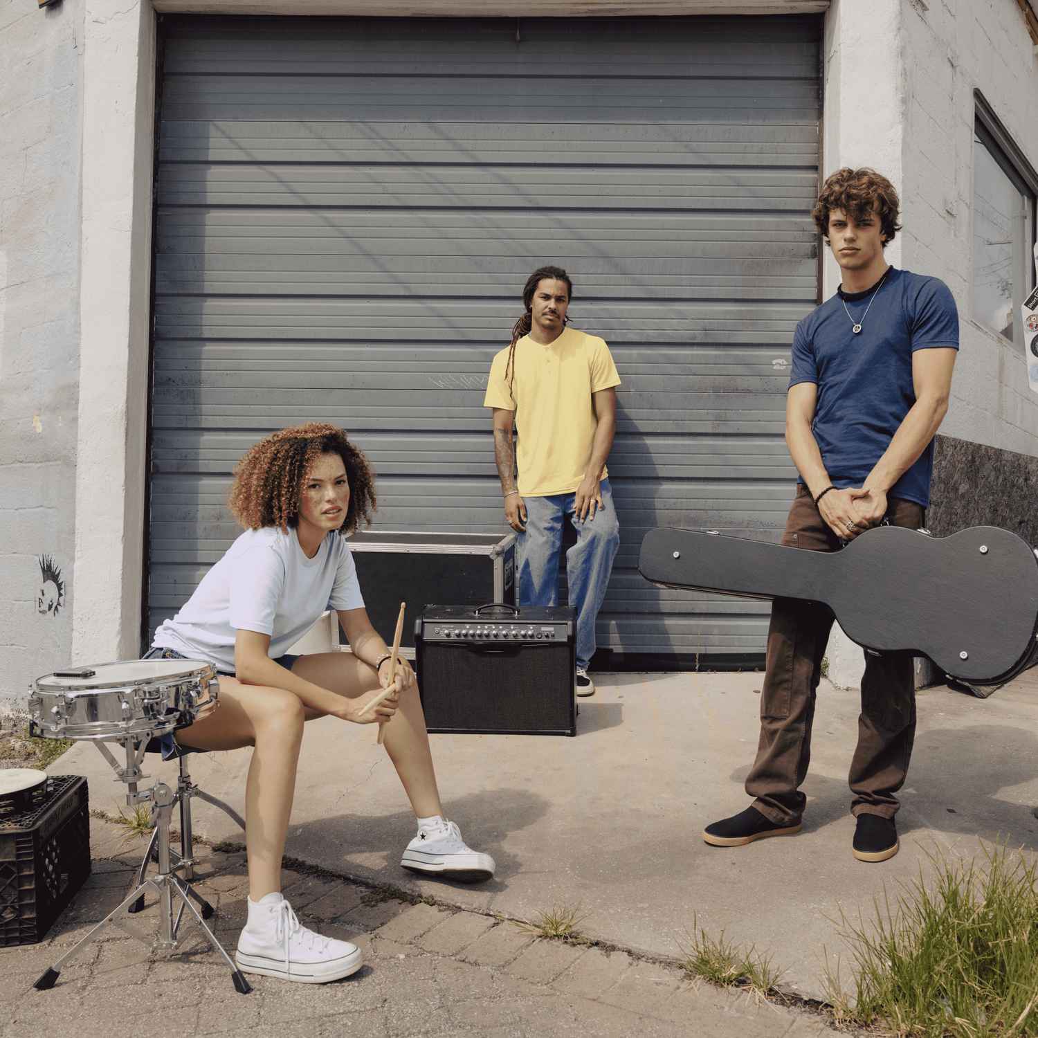 Deux hommes et une femme sont dehors devant un garage avec des instruments de musique.