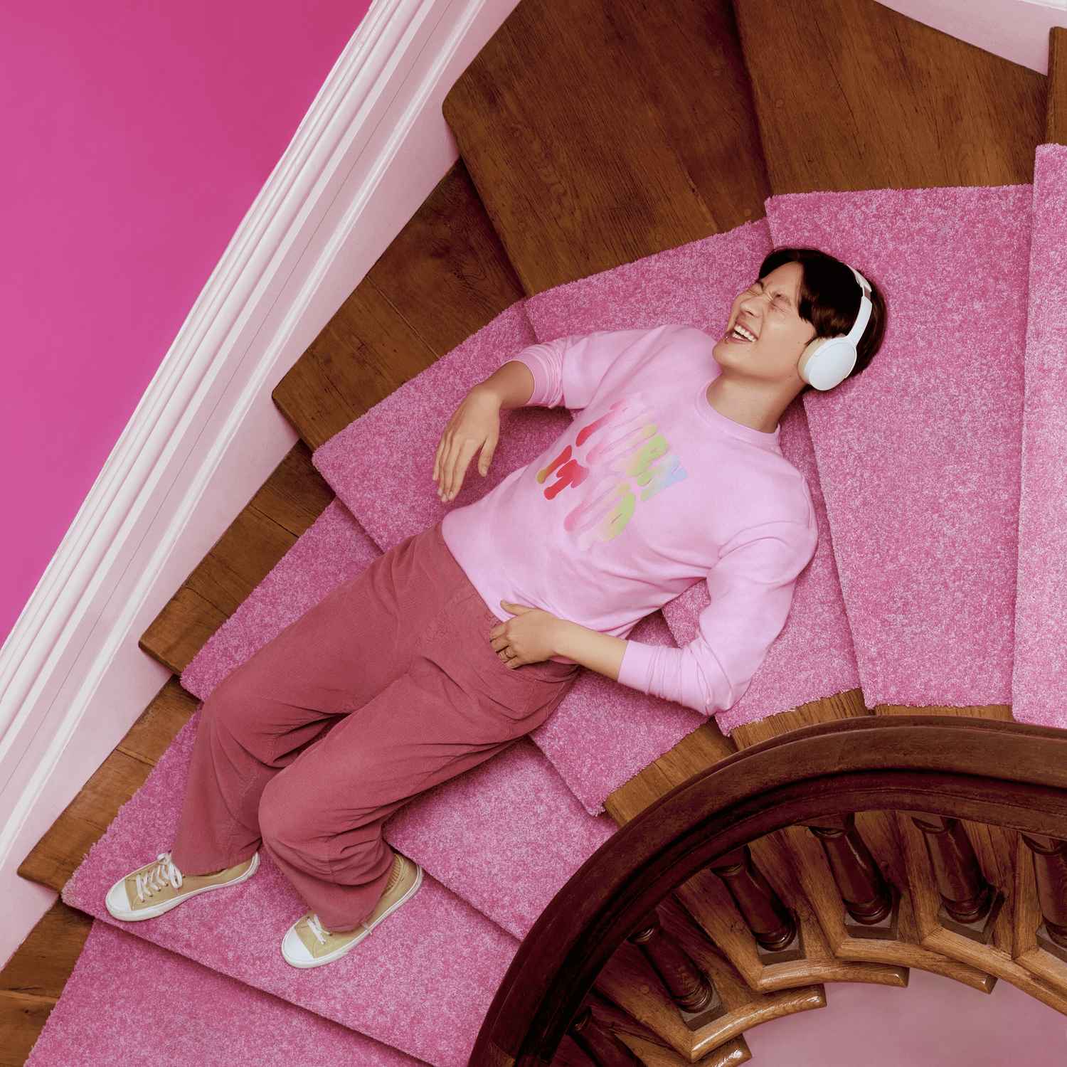 Une personne portant un tee-shirt Comfort Colors rose allongée sur un escalier, portant des écouteurs blancs et riant.