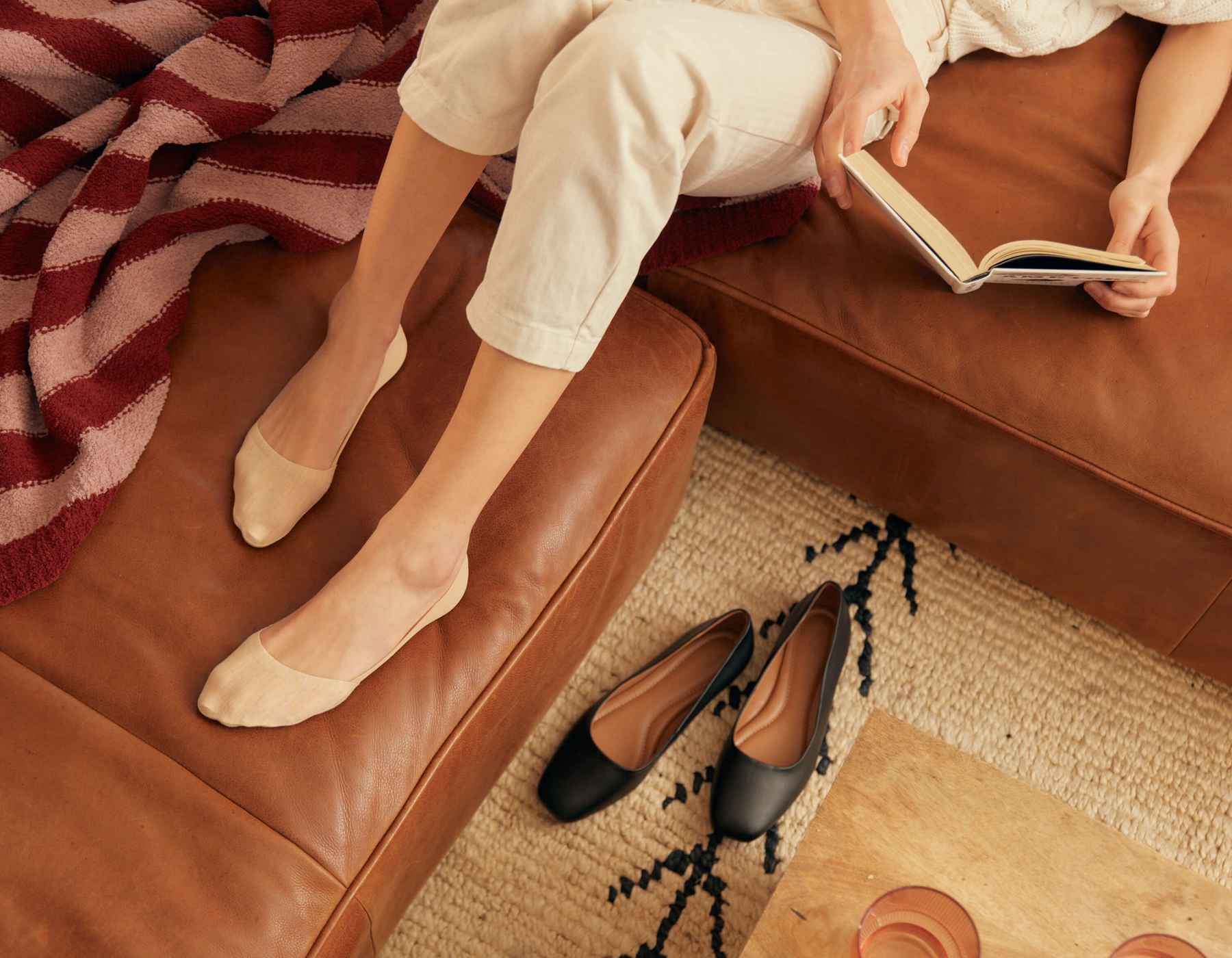 Une femme assise sur un canapé en cuir marron, portant des chaussettes Peds no show Peds, et lisant
