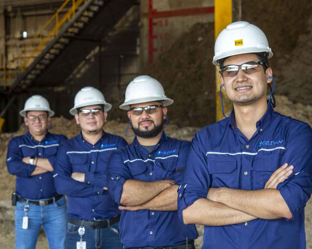 Quatre employés masculins portant des chemises Gildan et des casques de protection sourient.