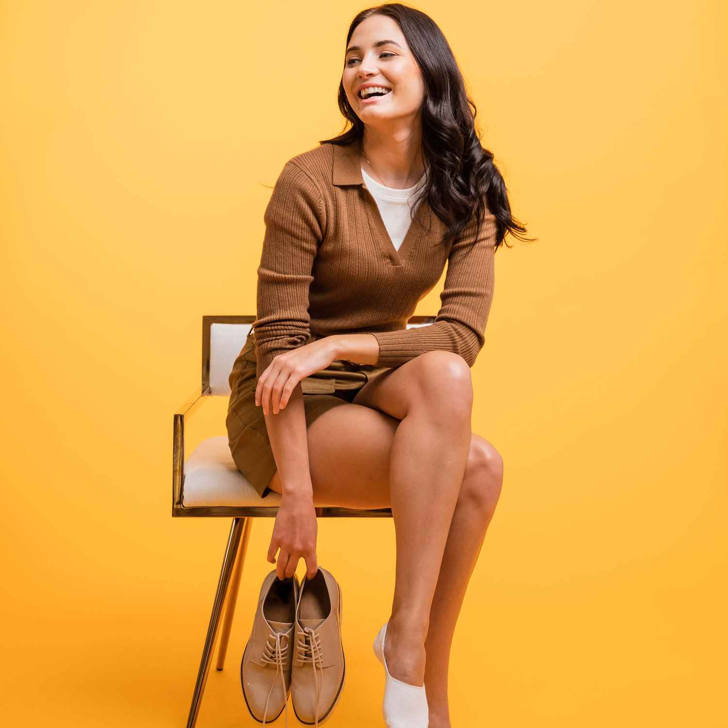 A woman is laughing while sitting on a chair and holding her shoes.